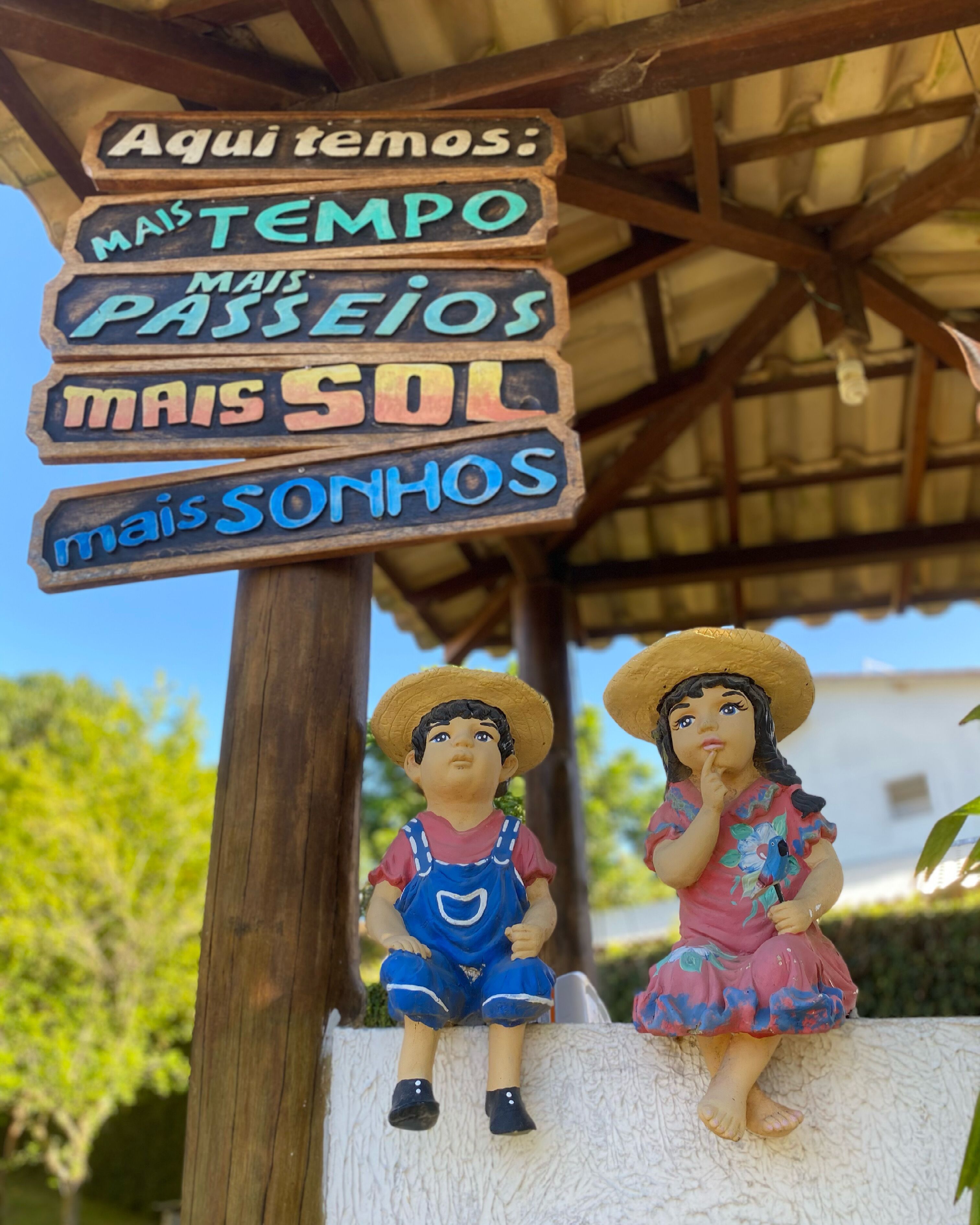 Un letrero de madera con texto en portugués, dos figuritas de niños y una estructura con techo de paja.