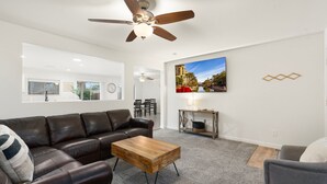 A living room with a brown leather sofa, a wooden coffee table, a ceiling fan, a TV mounted on the wall, and a fireplace. - Sunny Surprise Escape|Pool • Putting Green • Pets (Surprise)