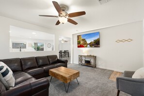 Ein Wohnzimmer mit braunem Ledersofa, einem Holztisch, einem Deckenventilator, einem an der Wand montierten Fernseher und einem Kamin.