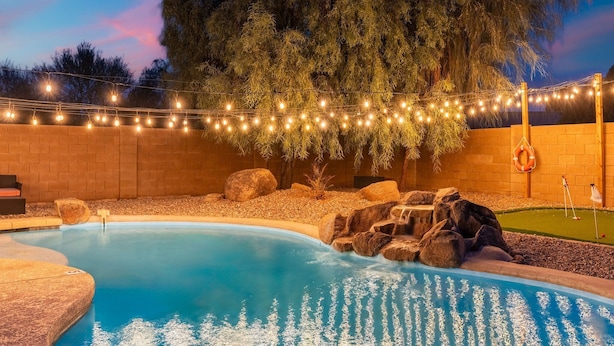 A swimming pool with a rock feature, surrounded by a brick wall and string lights. - Sunny Surprise Escape|Pool • Putting Green • Pets (Surprise)