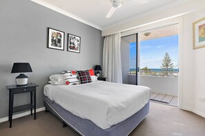 A bedroom with a bed, two bedside tables, a ceiling fan, a large window with a view of the sea, and two framed pictures on the wall. - Miss A l e x l Beachfront Babe l Pool & Parking! (Alexandra Headland)