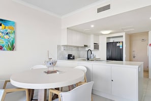 A modern kitchen with white cabinetry, a round dining table, and a small island with a sink. - Miss A l e x l Beachfront Babe l Pool & Parking! (Alexandra Headland)
