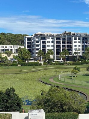 A modern multi-story building with a flat roof, surrounded by a well-maintained lawn and a small water feature. - Miss A l e x l Beachfront Babe l Pool & Parking! (Alexandra Headland)
