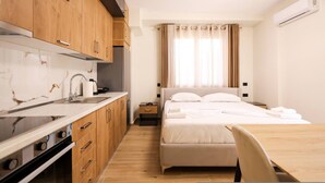 Ein kleines Hotelzimmer mit Küchenzeile, einem Bett mit weißer Bettwäsche, einem Holzesstisch und einem Fenster mit Vorhängen.