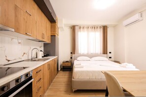 Ein kleines Hotelzimmer mit Küchenzeile, einem Bett mit weißer Bettwäsche, einem Holzesstisch und einem Fenster mit Vorhängen.