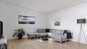 Ein modernes Wohnzimmer mit einem Ecksofa, einem Couchtisch, einer Stehlampe und einem großen Fenster mit Blick auf ein Haus.