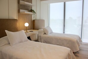 Ein Hotelzimmer mit zwei Betten, einem Nachttisch mit Lampe, einem Sessel und Blick auf die Stadt durch große Fenster.