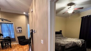 Quarto com cama, ventilador de teto, escrivaninha com cadeira e área de jantar com mesa e cadeiras.