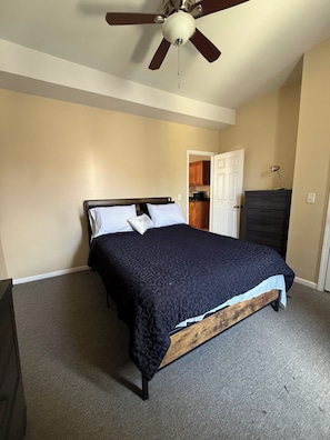 Un dormitorio con cama, ventilador de techo, cómoda y puerta que da a una cocina.