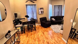 Uma sala de estar ampla com piso de madeira, uma mesa de jantar com cadeiras, um sofá preto e um ventilador de teto.