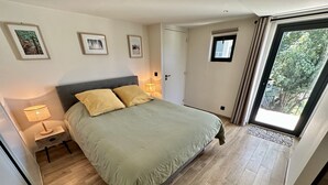 Un dormitorio con cama, lámparas de noche, cuadros enmarcados en la pared, una puerta y una ventana con vistas a follaje.