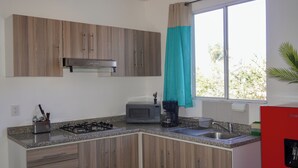 A kitchen with wooden cabinets, a microwave, a gas stove, a sink, and a window with a curtain. - Rest Between Matches | Cozy Depa (Tesistán)