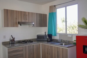 Una cocina con armarios de madera, microondas, estufa a gas, lavamanos y ventana con cortina.