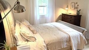 Un dormitorio con cama, lámpara de noche, ventana con cortinas, cómoda y una planta pequeña.