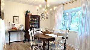 Comedor con mesa de madera y sillas blancas, una lámpara de araña y un ventanal con cortinas.