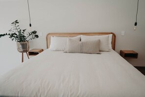 Una cama con colchón blanco, dos almohadas, cabecera trenzada, mesitas de noche y una planta colgante.