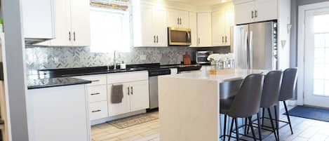 A modern kitchen with white cabinets, a central island with bar stools, stainless steel appliances, and a herringbone tile backsplash.