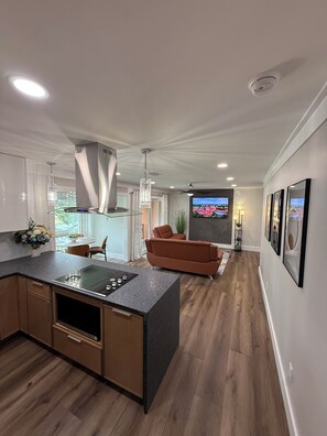 Eine moderne Küche mit Kochinsel, ein Essbereich mit Tisch und Stühlen sowie ein Wohnzimmer mit Sofa und Fernseher an der Wand.