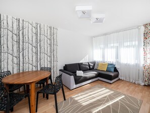 Ein Wohnzimmer mit einer Couch, einem Esstisch mit Stühlen, einer Wand mit Muster und einem Fenster mit durchsichtigen Vorhängen.