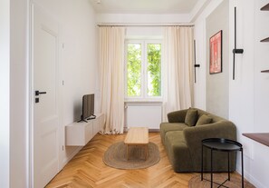 Ein Zimmer mit Holzboden, einem grünen Sofa, einem kleinen Holztisch, einem Fernseher auf einem Schrank und einem Fenster mit Vorhängen.