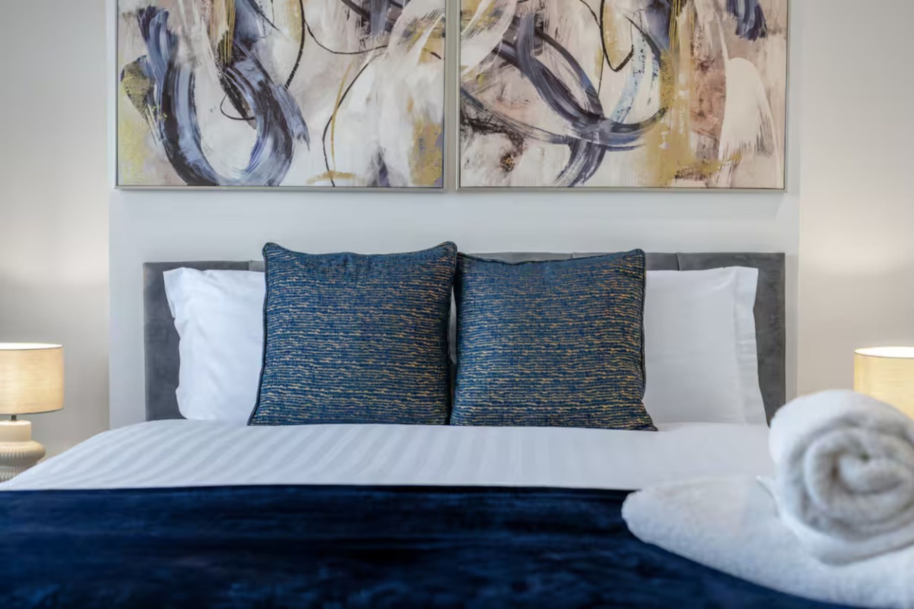 A hotel room with a bed, two abstract paintings, and a rolled towel.