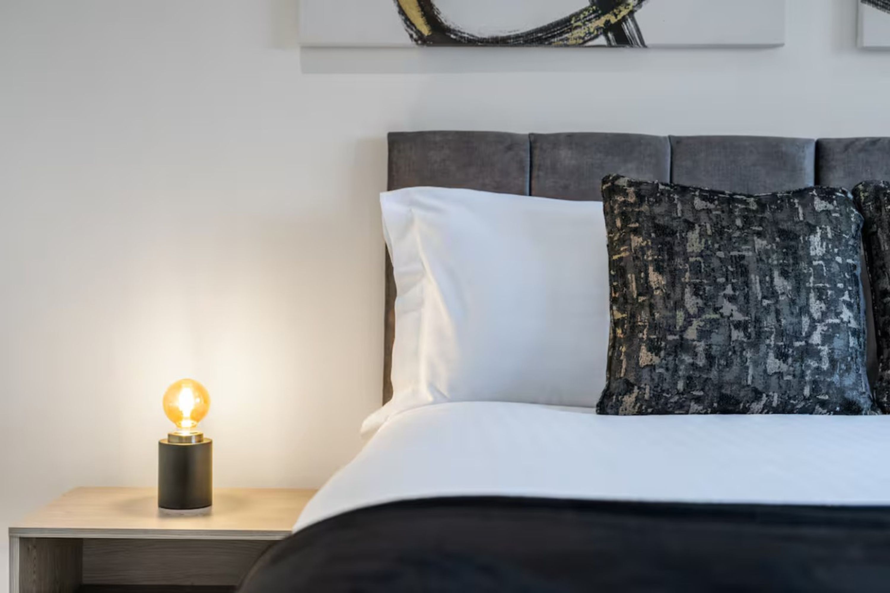 A bed with white and dark pillows, a nightstand with a lit bulb, and a wall with artwork.