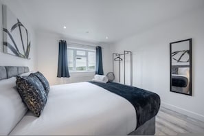 Una habitación de hotel moderna con una cama grande, una ventana con cortinas, un radiador, un perchero en la pared y una obra de arte abstracta enmarcada.