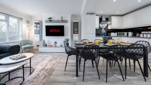 Et moderne køkken med hvide skabe, et spiseområde med sorte stole, et fladskærms-tv der viser 'NETFLIX' og en opholdsstue med sofa og sofabord.