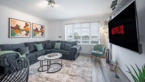 En moderne stue med en hjørnesofa, et sofabord, et fjernsyn ophængt på væggen og et vindue med persienner.