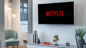 En moderne stue med et ophængt TV, der viser 'NETFLIX', en lyseblå lænestol, et sofabord i glas med dekorative genstande og en gulvlampe.