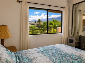 Et hotelværelse med en seng, et vindue med udsigt til palmer og huse, en lampe og en flytbar aircondition.