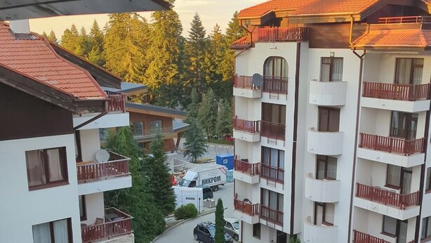 Un edificio residencial de varios pisos con balcones, un camión estacionado y árboles circundantes.