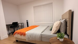 Ein Schlafzimmer mit einem Bett, einem Nachttisch mit Pflanze, einem Schreibtisch mit Stuhl und einem Fenster mit Jalousien.