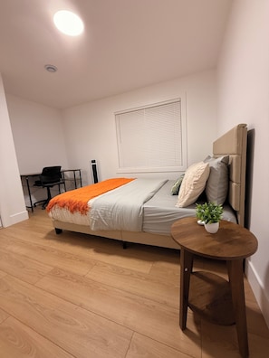 A bedroom with a bed, a bedside table with a plant, a desk with a chair, and a window with blinds.