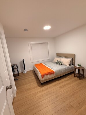 A bedroom with a bed, a bedside table, a window with blinds, a standing heater, and a door.