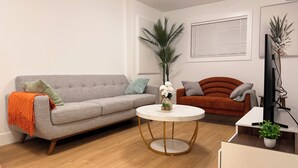 Een woonkamer met een grijze bank, een oranje plaid, een ronde salontafel, een bruine bank en een tv op een dressoir.