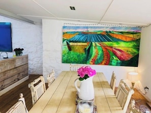 Una zona de menjador amb una taula de fusta llarga, cadires, un ram de flors en un vas i un quadre gran i colorit a la paret.
