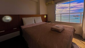 Ein Hotelzimmer mit Bett, Nachttisch, einer großen Fensterfront und Blick auf das Meer und Schiffe.