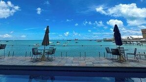 Blick über den Pool mit Liegestühlen und Sonnenschirmen, strahlend blauer Himmel und im Hintergrund Boote sowie Gebäude.