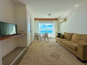 A living room with a television, a sofa, a dining table with chairs, and a view of the ocean through a large window. - Apartment for 6 People - Oceanfront (Fortaleza)