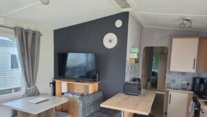 A compact RV interior with a kitchenette, a dining area with a table and bench, a TV mounted on a stand, and a small trash bin. - Seaside Haven Retreat in Skegness (Skegness)