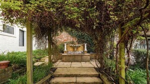 A stone pathway leading to a stone bench with a decorative top, surrounded by greenery and a brick wall. - Villa Sleeps up to 17 - Hot Tub, Garden, Parking (Pinner)