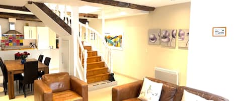 A living room with a brown leather sofa, a coffee table, and a dining area with a wooden table and chairs. A staircase leads to an upper level.