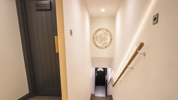 En hall med trägolv, en mörk dörr med skylten ”Lägenhet 2” och en lampa med ett geometriskt mönster på väggen.