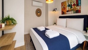 Ein Hotelzimmer mit Bett, Nachttisch, Wand-Klimaanlage, dekorativem Spiegel und einem farbenfrohen abstrakten Gemälde.