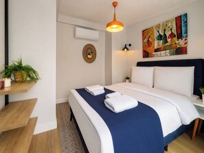 Ein Hotelzimmer mit Bett, Nachttisch, Wand-Klimaanlage, dekorativem Spiegel und einem farbenfrohen abstrakten Gemälde.