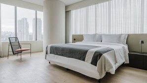 Quarto de hotel moderno com uma cama grande, uma poltrona, mesa de cabeceira e vista para a paisagem urbana.