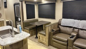 A compact RV interior with a kitchenette, a seating area, and a window with blinds.