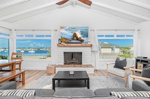 Ein geräumiges Wohnzimmer mit Kamin, einem großen Gemälde einer Küstenlandschaft und Blick auf Meer und Boote durch große Fenster.