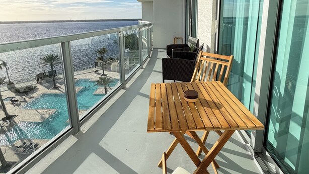 En balkon med et træbord og stole, med udsigt over en pool og havet.